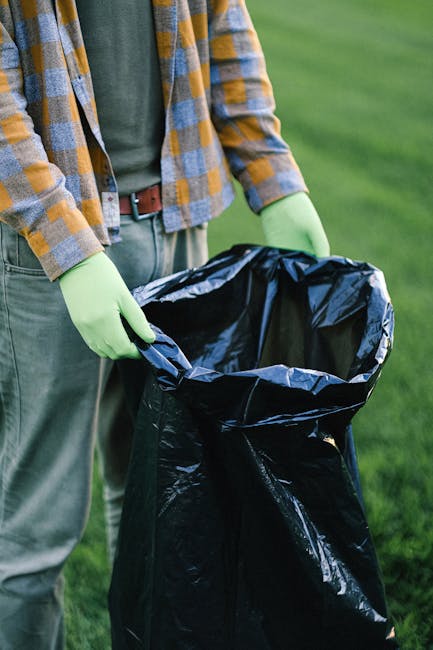 A large pile of mixed waste including black and red garbage bags, flattened cardboard boxes, and various paper and plastic packaging, is piled outside a row of waste bins on a paved sidewalk in an urb